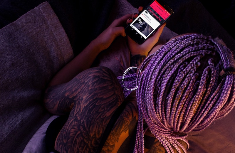 Girl with braids, sitting in dark room lit with red while looking at cell phone.