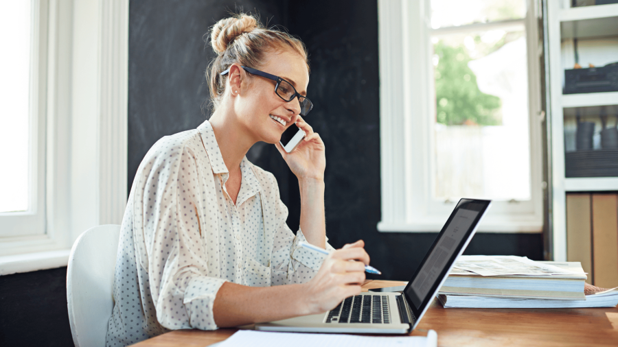 A woman working from home on her laptop talking on her cellphone.