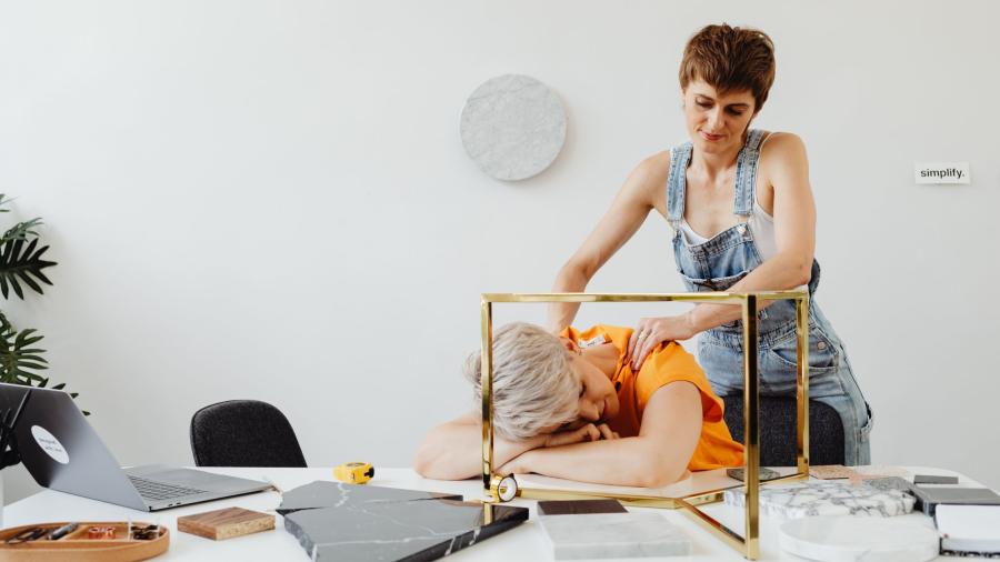 A women massaging another person's shoulders as they lay their head down on their messy desk. 