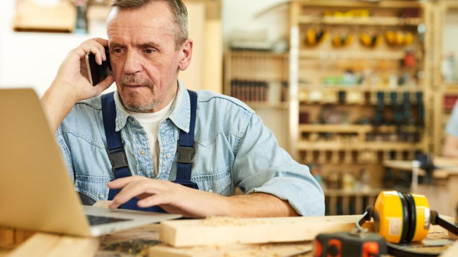 A man sitting on a the phone looking at his laptop in his workshop.